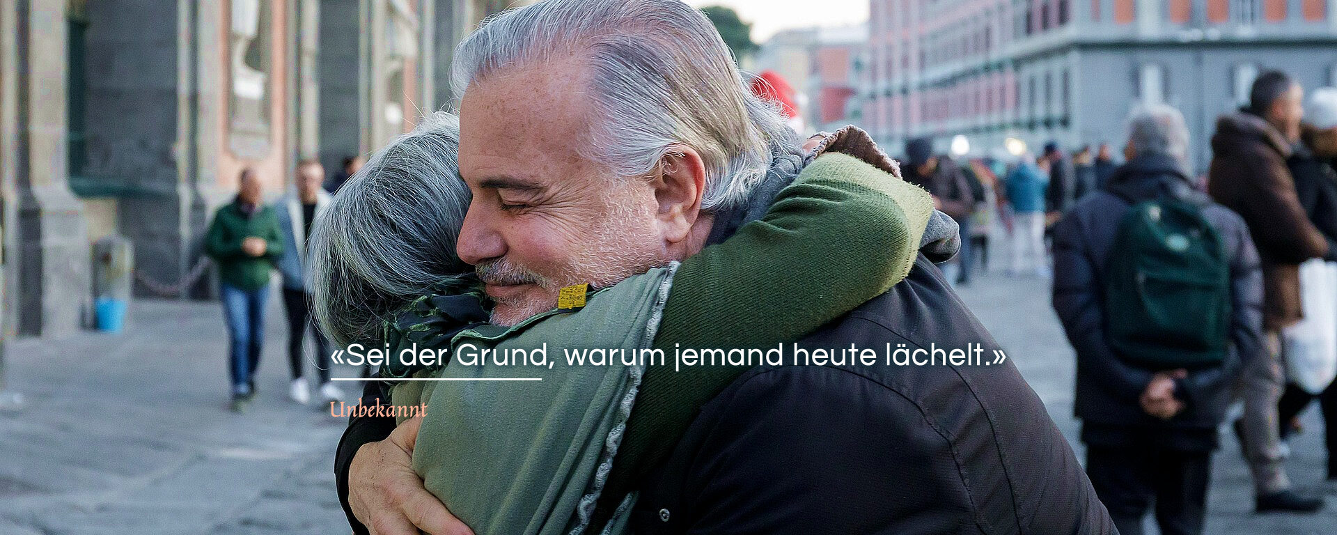 Auf einem Platz mit historischen Gebäuden, umarmt ein älterer Mann eine Frau und lächelt dabei berührt